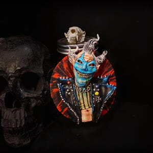Puede incluir: Una escultura detallada de un payaso con piel azul, ojos rojos y una sonrisa amenazante. El payaso lleva una chaqueta de cuero negra con tachuelas plateadas y una cadena. También se ven una calavera y una calavera de gato.