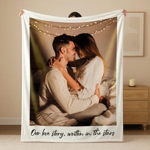 May include: A white fleece blanket featuring a photograph of a couple embracing. The image is framed with a white border and includes the text "Our love story, written in the stars" at the bottom. Fairy lights are visible in the background.