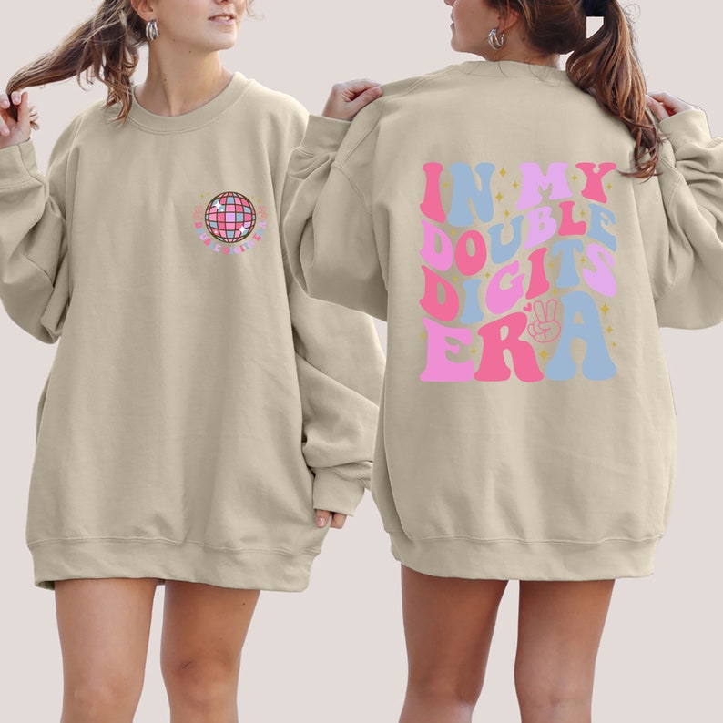 May include: A beige sweatshirt with a disco ball graphic on the front and the text "In My Double Digits Era" on the back. The text is in pink, blue, and gold.
