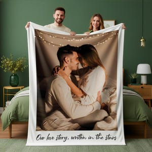 May include: A white blanket featuring a photograph of a couple embracing, with string lights in the background. The text "Our love story, written in the stars" is printed at the bottom. Two people are holding the blanket.