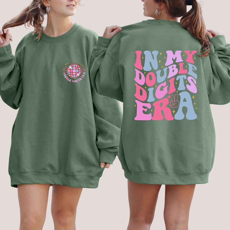 May include: Two green sweatshirts with different designs. The front sweatshirt has a disco ball graphic with the text "In My Double Digits Era". The back sweatshirt has the text "In My Double Digits Era" in a retro font with pink, blue, and white colors.