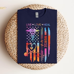 May include: A navy blue t-shirt with a rainbow-colored American flag design. The flag features a stethoscope, a heart, and the words "Live Love Heal Nurse".