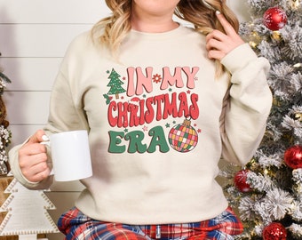 May include: A cream-colored sweatshirt with the text "IN MY CHRISTMAS ERA" in red, green, and pink. The design includes a Christmas tree, a flower, stars, and a disco ball ornament. The sweatshirt is a festive holiday apparel item.