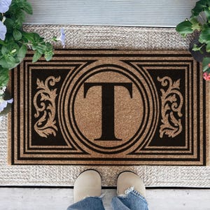 May include: A rectangular coir doormat featuring a natural tan fiber background with a bold black border and a central black circle containing the letter T. The design includes symmetrical black scrollwork patterns on the left and right sides.