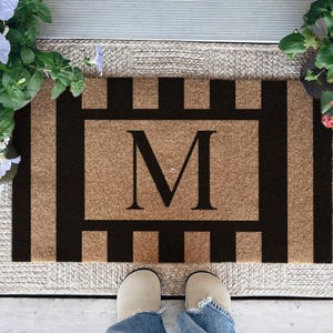 May include: A rectangular doormat with a brown background and black vertical stripes. The center features a large black letter "M". The mat is surrounded by a woven border and is placed on a wooden porch. Potted flowers in bloom are on either side.