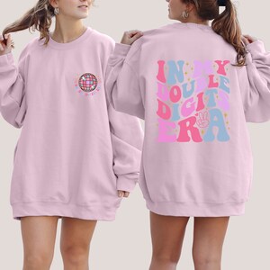 May include: Two pink sweatshirts with different designs. The front sweatshirt has a disco ball graphic with the text "Disco til Dawn". The back sweatshirt has the text "In My Double Digits Era" in a retro font with stars and a peace sign.