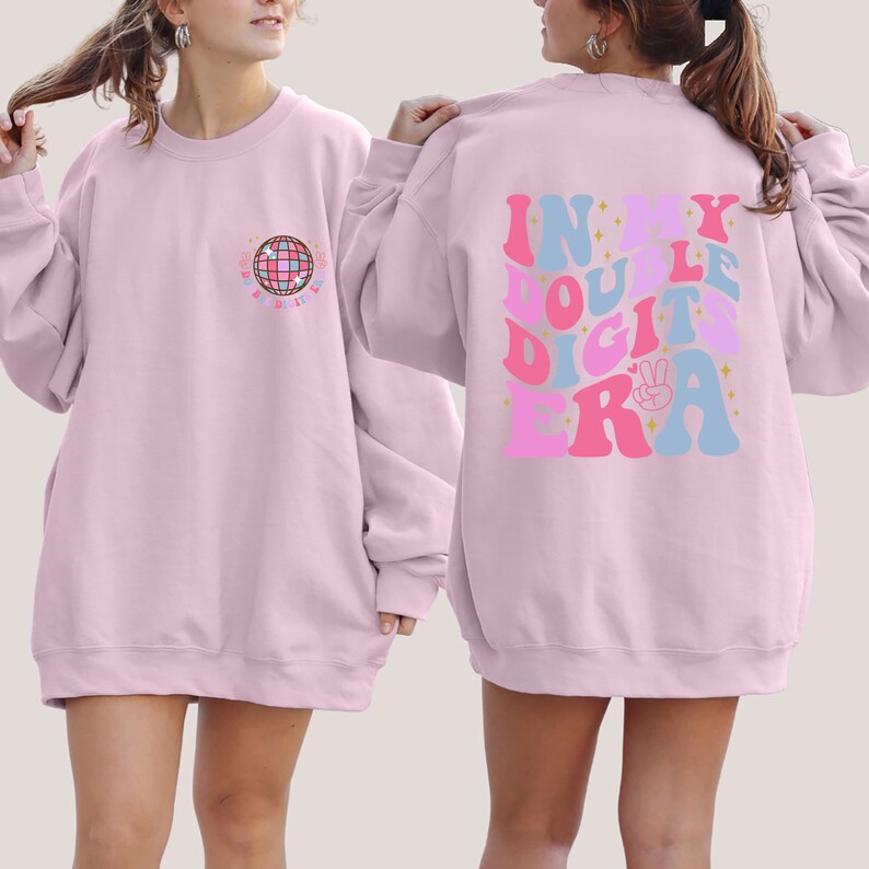 May include: Two pink sweatshirts with different designs. The front sweatshirt has a disco ball graphic with the text "Disco til Dawn". The back sweatshirt has the text "In My Double Digits Era" in a retro font with stars and a peace sign.