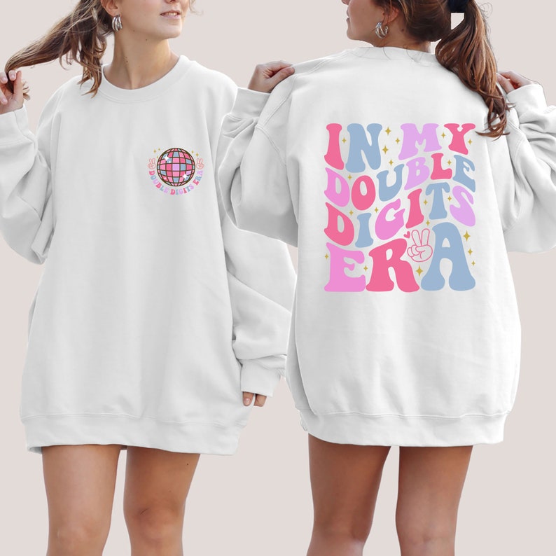 May include: White sweatshirt with a disco ball graphic on the front and the text "In My Double Digits Era" on the back. The text is in pink, blue, and white with a peace sign and glitter accents.
