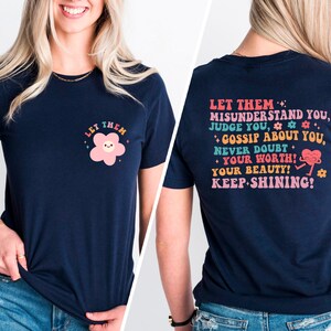May include: Navy blue t-shirt with a pink flower graphic on the front and a motivational message on the back. The message reads "Let them misunderstand you, judge you, gossip about you, never doubt your worth! Your beauty! Keep shining!"
