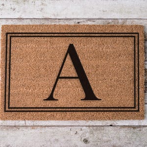 May include: A rectangular tan coir doormat featuring a large, centered black capital letter A. The letter is enclosed within a double-line black rectangular border that frames the perimeter of the mat, creating a clean and structured appearance.