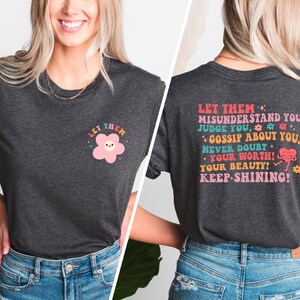 May include: A dark gray t-shirt with a pink flower graphic on the front and the text "LET THEM MISUNDERSTAND YOU, JUDGE YOU, GOSSIP ABOUT YOU, NEVER DOUBT YOUR WORTH! YOUR BEAUTY! KEEP SHINING!" on the back.