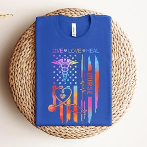 May include: A blue t-shirt with a rainbow-colored American flag design. The flag features a caduceus, a heart, and the text "Live Love Heal Nurse".