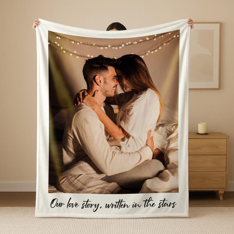 May include: A white fleece blanket featuring a photograph of a couple embracing. The image is framed with a white border and includes the text "Our love story, written in the stars" at the bottom. Fairy lights are visible in the background.