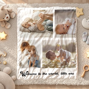 May include: A baby photo blanket with four family photos, surrounded by baby toys and accessories. The blanket has the text "Welcome to the world, little one" in black script. The toys include a teddy bear, star-shaped lights, and wooden toys.