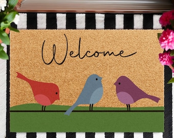 May include: A rectangular doormat with a brown background and the word "Welcome" in black script. Three colorful birds perch on a green line. The mat is on a black and white striped surface.