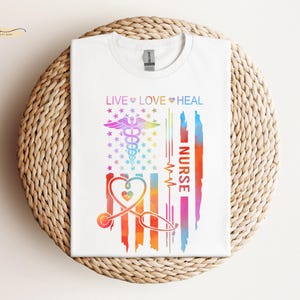 May include: White t-shirt with a rainbow-colored American flag design. The flag has a stethoscope, a heart, and the words "Live Love Heal Nurse".