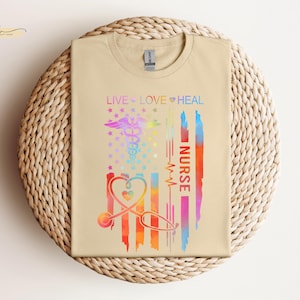 May include: A beige t-shirt with a rainbow-colored American flag design. The flag has a stethoscope and the words "Live Love Heal Nurse" on it.