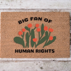 May include: A rectangular doormat made of natural coir fiber. The mat features a floral design with orange flowers and green leaves. The text "BIG FAN OF HUMAN RIGHTS" is printed in black across the top and bottom.