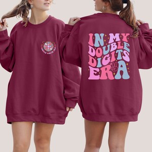May include: Burgundy sweatshirt with a disco ball graphic on the front and the text "In My Double Digits Era" on the back. The text is in pink, blue, and white.