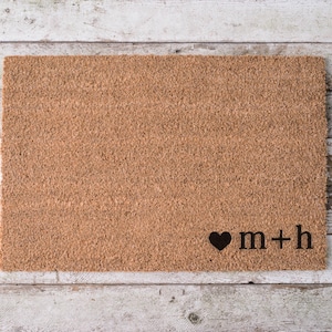 May include: A rectangular tan coir doormat with a coarse, fibrous texture. In the bottom right corner, there is a black printed design consisting of a solid heart symbol followed by the lowercase letters m, a plus sign, and the letter h.