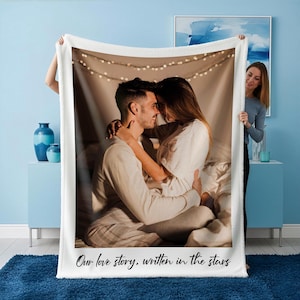 May include: A white fleece blanket featuring a printed photograph of a couple embracing. The image is framed by a white border, and the text "Our love story, written in the stars" is printed at the bottom. The blanket is held up by a person.