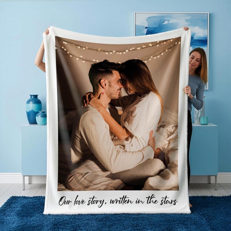 May include: A white fleece blanket featuring a printed photograph of a couple embracing. The image is framed by a white border, and the text "Our love story, written in the stars" is printed at the bottom. The blanket is held up by a person.