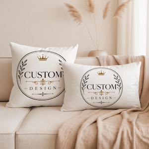 May include: Two white throw pillows featuring a circular black and gold emblem. The design includes a crown icon, laurel branches, and the text "CUSTOM DESIGN" centered within the circle. The pillows are square and rectangular in shape.