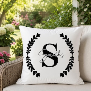 May include: A square white throw pillow featuring a black graphic design. The design consists of a large capital letter S centered within a circular laurel wreath made of leaves, with the word Smiths written in a cursive script across the letter.