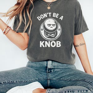 May include: A dark grey t-shirt with the white text "DON'T BE A KNOB". The graphic shows a grumpy-faced knob design. The shirt is worn with blue jeans and white trainers, creating a casual, everyday look.