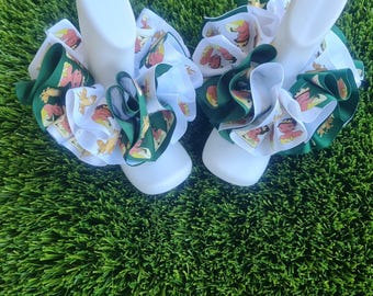 May include: A pair of ruffled ankle socks featuring a layered design of white fabric and green ribbon. The ribbon is decorated with a repeating graphic print of a cartoon character wearing a yellow dress and a crown, set against a green background.