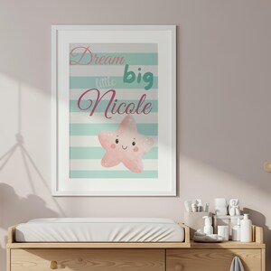 May include: A framed print with the text "Dream little big Nicole" and a pink star illustration. The print is above a wooden changing table. The room is decorated in neutral tones, with a hanging shelf and a small bin.