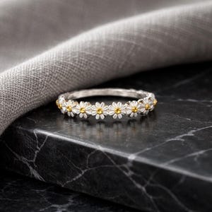 May include: A silver ring featuring a repeating daisy flower design. Each daisy has white petals and a gold-colored center. The ring is set against a dark, textured surface with a gray cloth in the background.