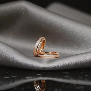 May include: Two gold rings, each with a band of small, clear gemstones. They rest on a grey, silky surface, with a reflection of the rings below. The rings are likely wedding or engagement rings.