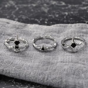以下が含まれることがあります： 黒い宝石がアクセントになった3つのシルバーリング。星と枝のようなデザインで、1つのリングには2つの宝石が付いています。リングはグレーの布の上に展示されています。