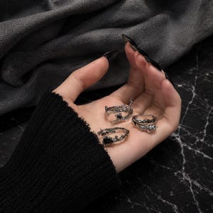 Puede incluir: Cuatro anillos plateados con diseños intrincados y texturizados. Los anillos varían en estilo, algunos con detalles oscuros y otros con una apariencia más abierta y ornamentada. Los anillos se presentan sobre un fondo oscuro.