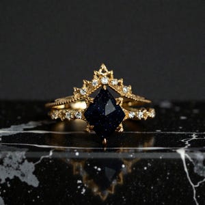 Op de afbeelding: Een gouden ringenset met een donkerblauwe, geometrisch geslepen edelsteen. De ring is versierd met kleine, heldere edelstenen en ingewikkelde details. De set bevat een kroonvormige band en een bijpassende band.