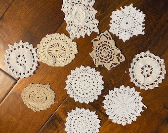 May include: A collection of handmade crochet doilies in various shapes and sizes. The doilies are primarily white and beige, with intricate lace patterns. Some feature floral designs, while others have geometric shapes. They are arranged on a wooden surface.
