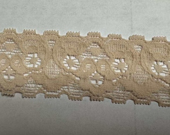 May include: Close-up of a tan lace trim with a scalloped edge. The lace features a repeating floral pattern with openwork details. This decorative trim is suitable for various craft and sewing projects.
