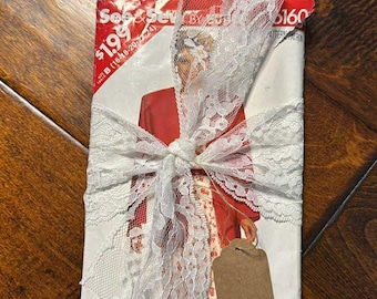 May include: A vintage sewing pattern packet tied with white lace ribbon. The pattern packet is red and white with the text "See & Sew" and the number "6160". A small brown tag is attached to the ribbon.