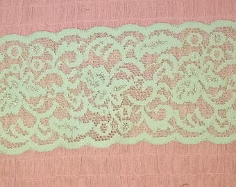 May include: A length of light green lace with a scalloped edge. The lace features a detailed floral pattern. The fabric is displayed against a pink background, highlighting the delicate design and color of the lace.