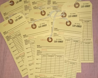 May include: A collection of vintage layaway tags in a fan shape. The tags are a light yellow color with printed text and lines for information. Each tag has a string attached for hanging.