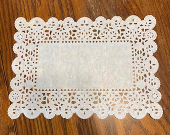 May include: White paper doily with a delicate lace pattern. The doily is square shaped and has a smooth, white surface.
