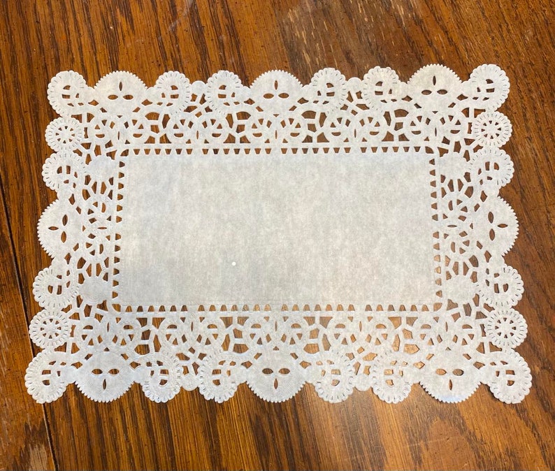 May include: White paper doily with a delicate lace pattern. The doily is square shaped and has a smooth, white surface.