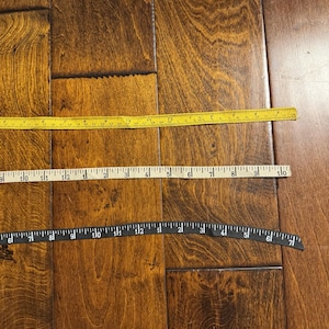 May include: Two measuring tapes, one yellow and one black, are laid out on a wooden floor. The yellow tape measures up to 16 inches and the black tape measures up to 7 inches.