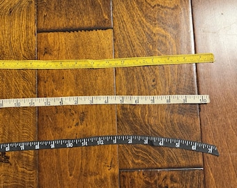May include: Two measuring tapes, one yellow and one black, are laid out on a wooden floor. The yellow tape measures up to 16 inches and the black tape measures up to 7 inches.