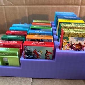May include: A purple tiered organizer holding a collection of colorful Little Tikes audio cassettes. The cassettes have various titles, including "Axel the Truck: Rocky Road" and "Careful Gamble!" The organizer is made of plastic.