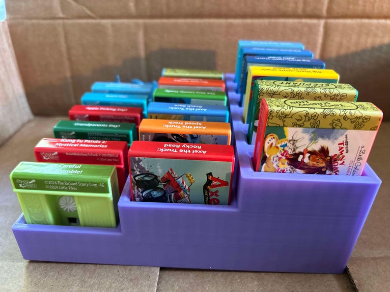 May include: A purple tiered organizer holding a collection of colorful Little Tikes audio cassettes. The cassettes have various titles, including "Axel the Truck: Rocky Road" and "Careful Gamble!" The organizer is made of plastic.