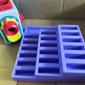 May include: A purple tiered display stand with multiple rectangular compartments. A colorful toy camera with red, yellow, blue, and white accents is in the background. The stand is designed to hold small items.