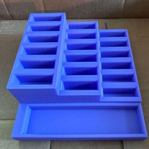 May include: A purple tiered display stand with multiple rectangular compartments. The stand has a stepped design, with three sections of varying heights, and a rectangular base. The stand is made of a solid material and is designed to hold small items.