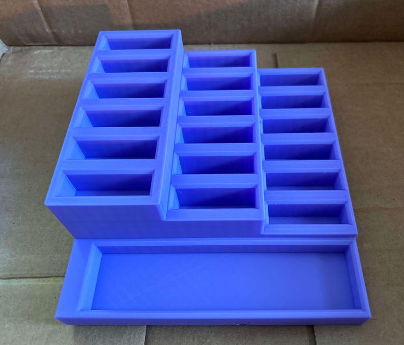 May include: A purple tiered display stand with multiple rectangular compartments. The stand has a stepped design, with three sections of varying heights, and a rectangular base. The stand is made of a solid material and is designed to hold small items.